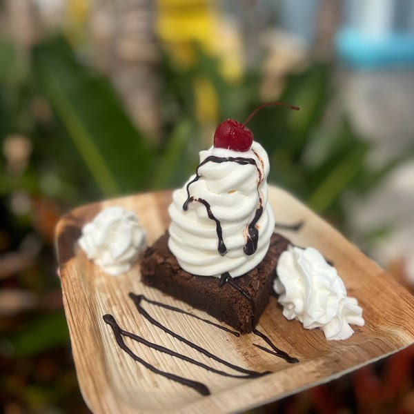 Brownie topped with whipped cream, chocolate drizzle, and a cherry on a wooden plate.