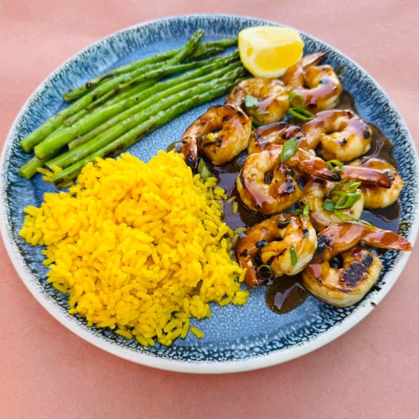 Grilled shrimp with dark sauce, yellow rice, asparagus, and a lemon wedge on a blue plate.