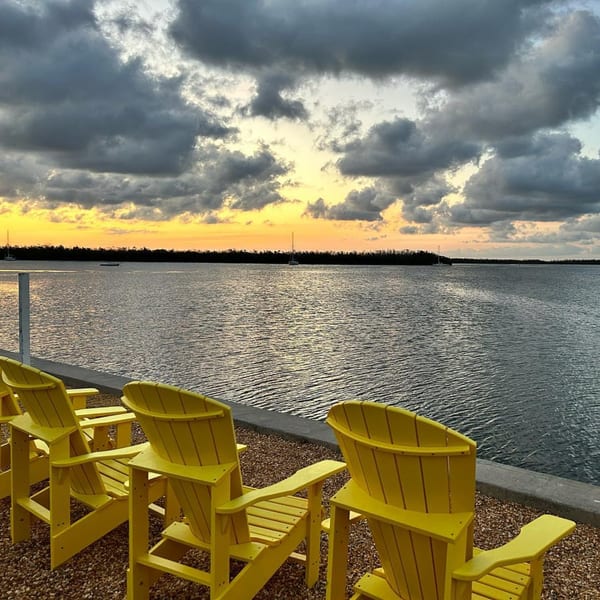 Yellow Adirondack chairs face a calm body of water under a dramatic sunset sky at Beachcomber Trading Post.