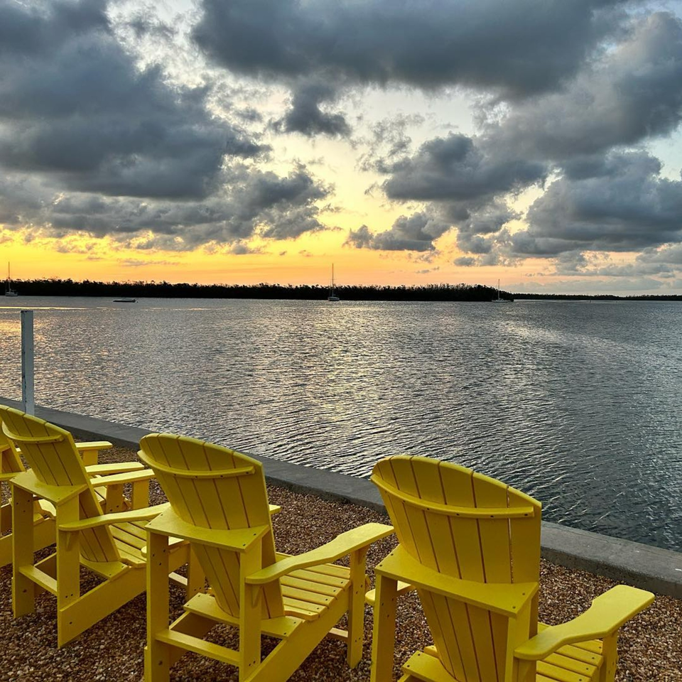Yellow Adirondack chairs face a calm body of water under a dramatic sunset sky at Beachcomber Trading Post.