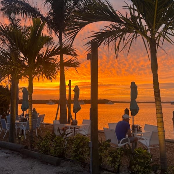 A vibrant orange sunset over the water, with palm trees and outdoor dining tables at Beachcomber Trading Post.
