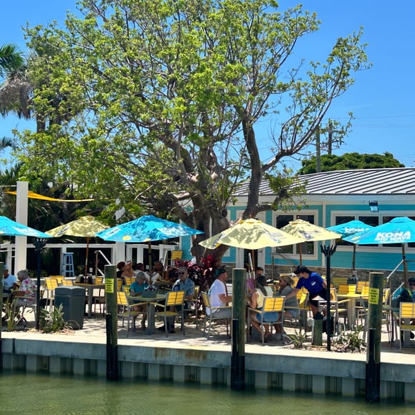 Outdoor dining area at Beachcomber Trading Post Restaurant with patrons seated under colorful umbrellas by the water.