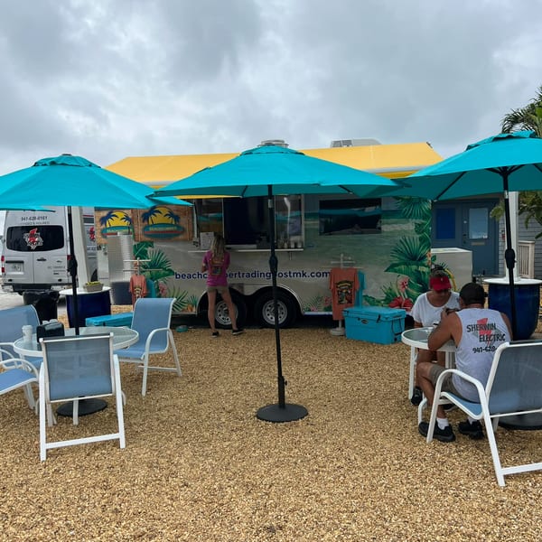 Outdoor seating area with turquoise umbrellas and a Beachcomber Trading Post food truck.