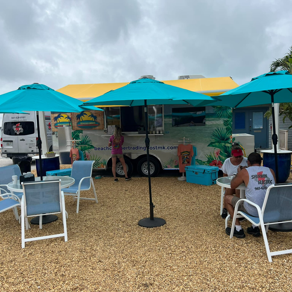 Outdoor seating area with turquoise umbrellas and a Beachcomber Trading Post food truck.