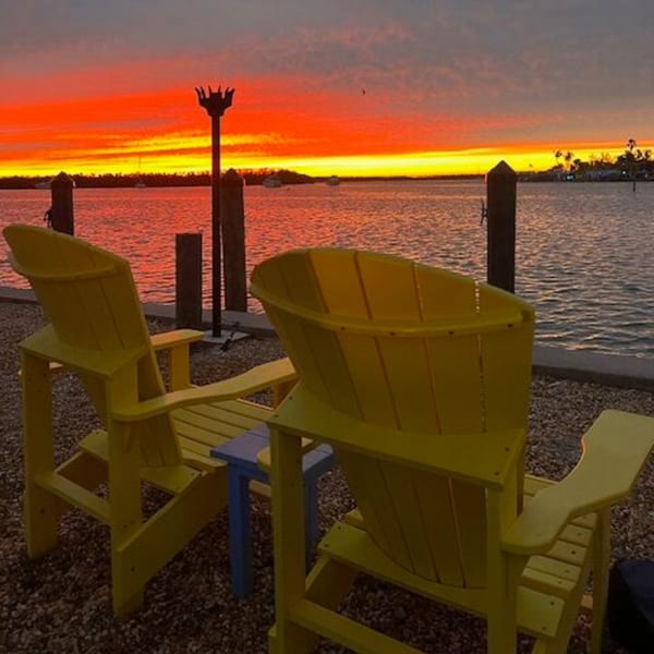 Two yellow Adirondack chairs face a vibrant orange and red sunset over the water at Beachcomber Trading Post.