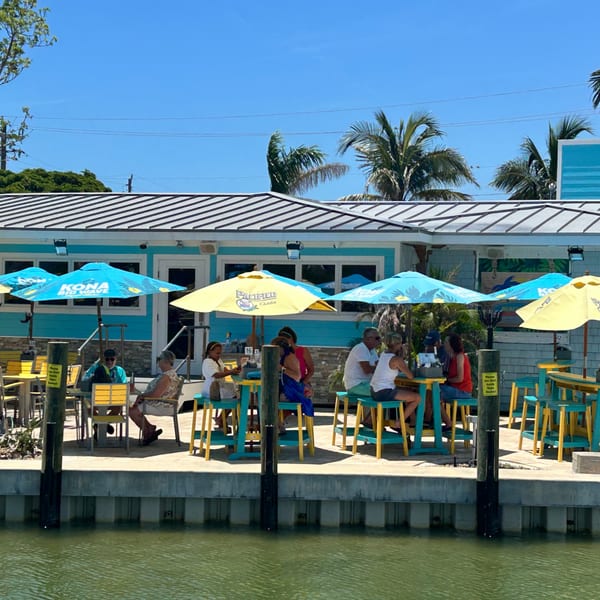 Diners enjoy outdoor seating under blue and yellow umbrellas at Beachcomber Trading Post Restaurant, overlooking the water.