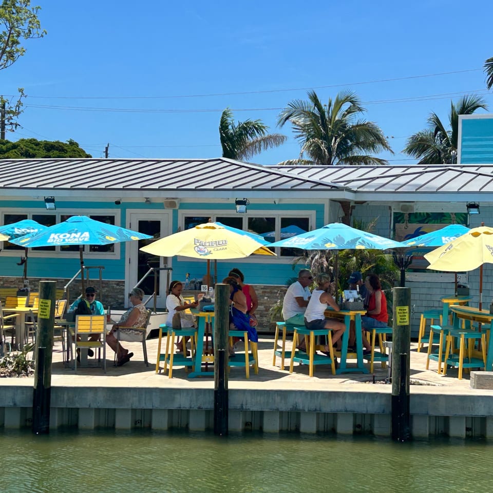 Diners enjoy outdoor seating under blue and yellow umbrellas at Beachcomber Trading Post Restaurant, overlooking the water.