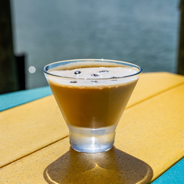 Espresso martini with coffee beans on top, on a yellow and blue table with water in the background.