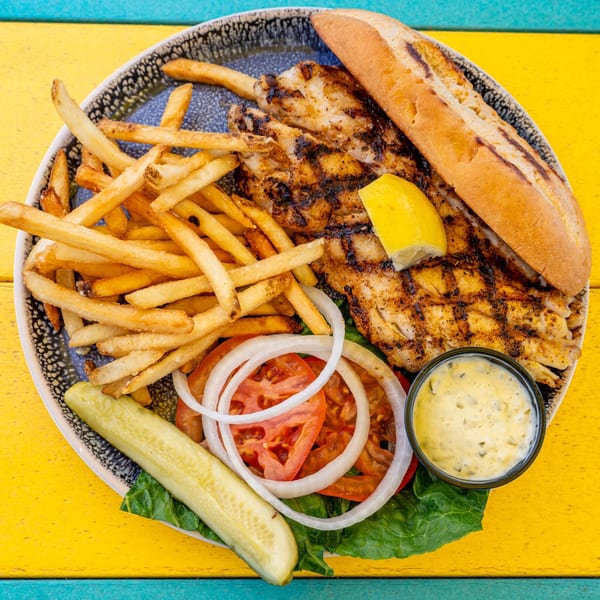 Grilled fish sandwich with fries, lettuce, tomato, onion, pickle, and tartar sauce on a colorful table.
