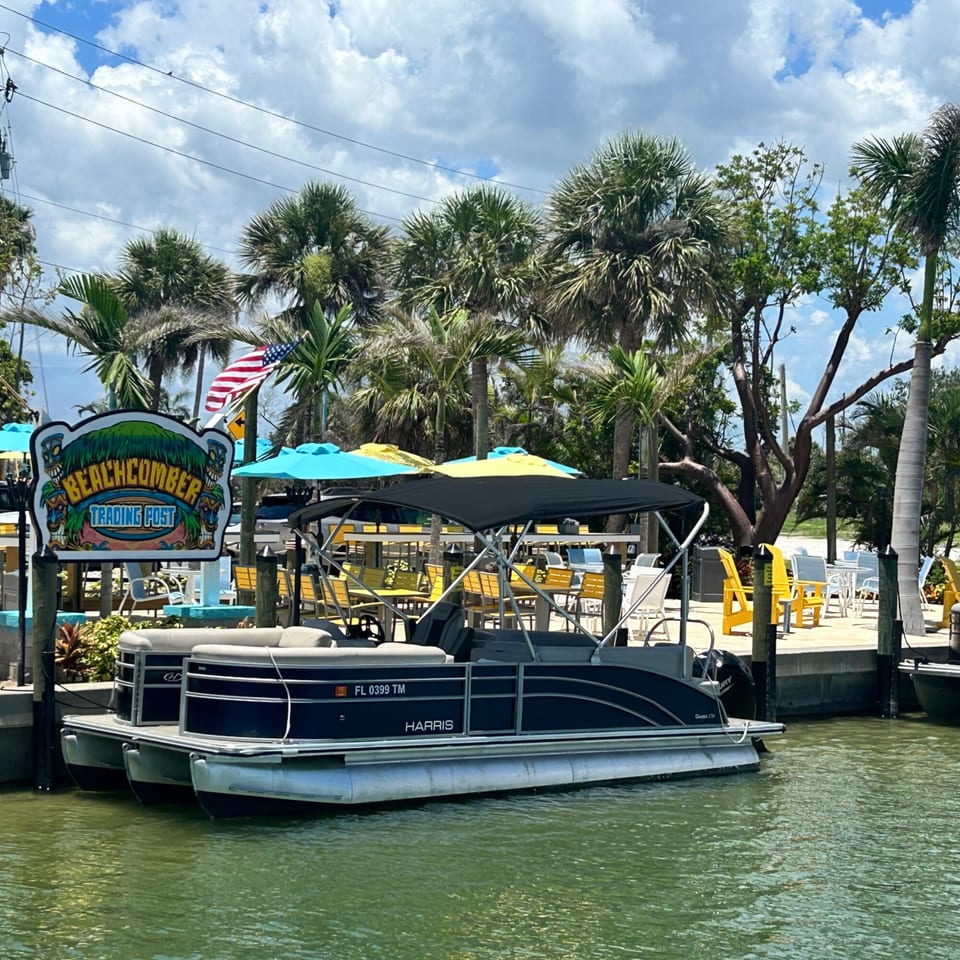 Beachcomber Trading Post sign, pontoon boat, and outdoor seating area with palm trees.