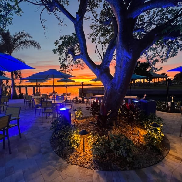 Outdoor dining patio at Beachcomber Trading Post Restaurant with blue lighting and a vibrant sunset over water.