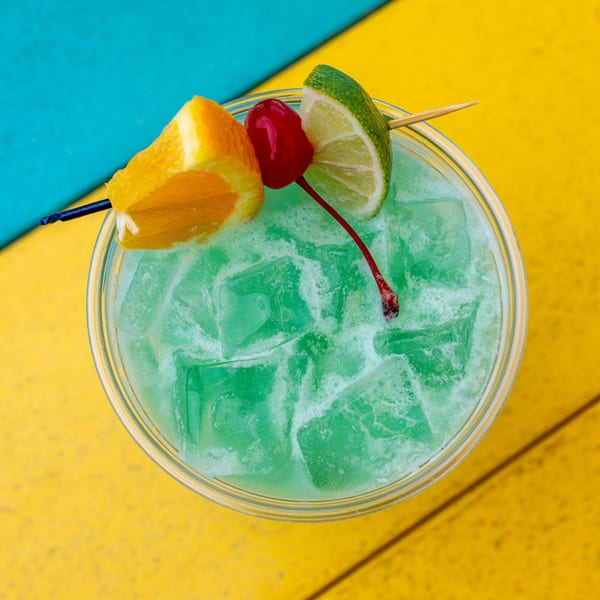 A vibrant green cocktail with ice, garnished with an orange slice, cherry, and lime wedge.