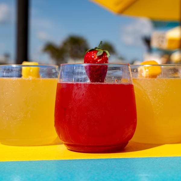 Three vibrant drinks, two yellow and one red with a strawberry garnish, on a colorful table.