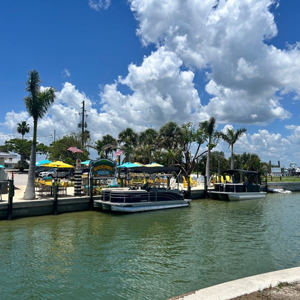 Beachcomber Trading Post Restaurant with outdoor seating, palm trees, and boats docked on a sunny day.