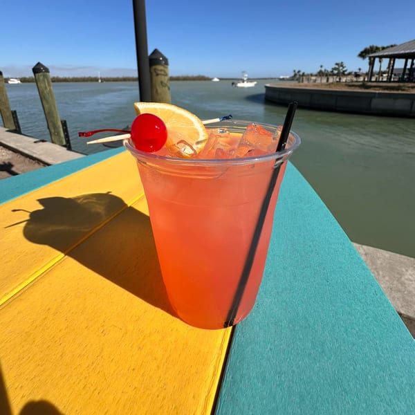 A pink cocktail with a cherry and orange slice sits on a colorful table by the water.