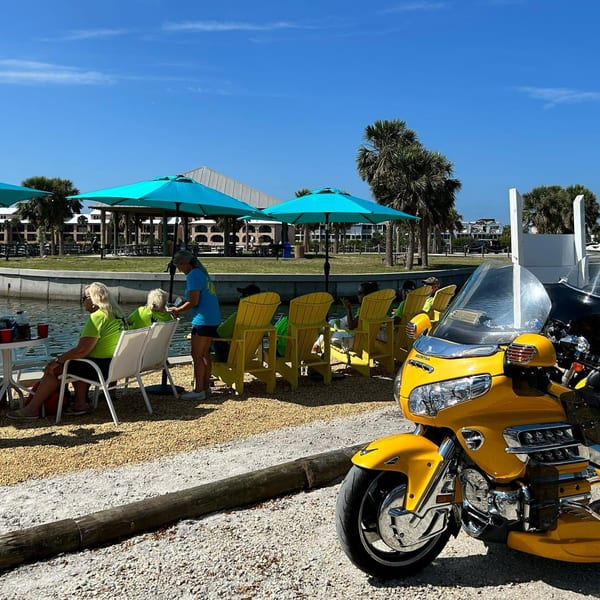 People dining outdoors by the water under blue umbrellas, with a yellow motorcycle in the foreground.