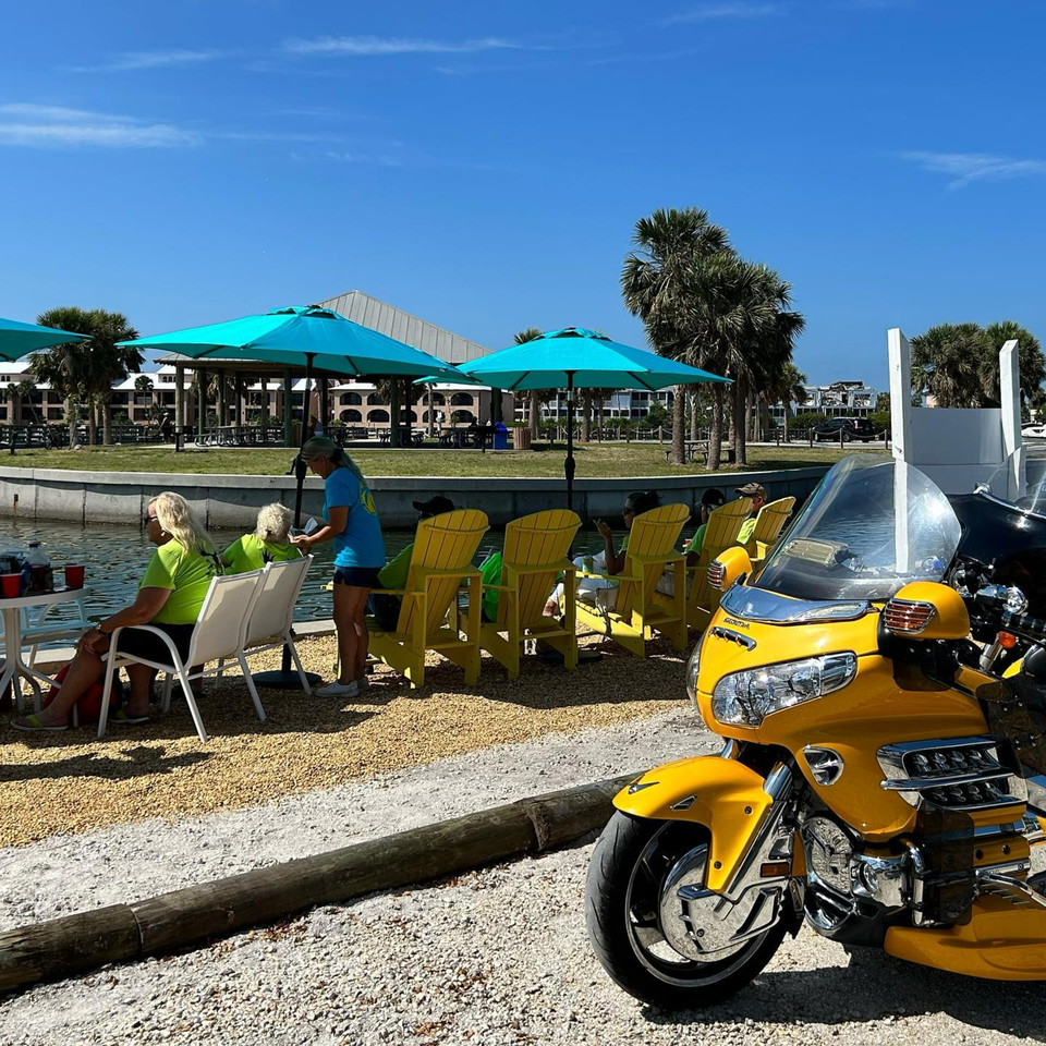 People dining outdoors by the water under blue umbrellas, with a yellow motorcycle in the foreground.