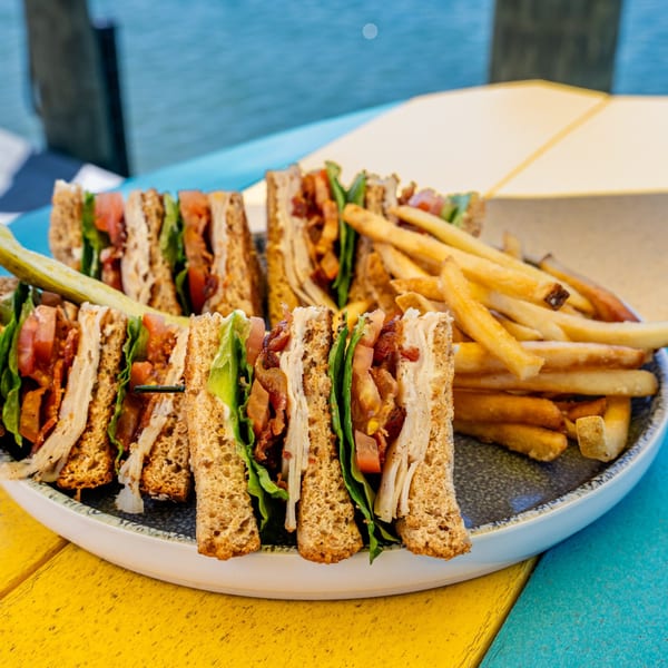 A club sandwich with fries and a pickle spear on a plate, on a colorful wooden table.