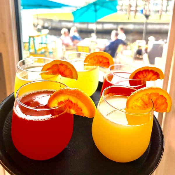 A tray of red and yellow cocktails garnished with orange slices, with a blurred outdoor patio in the background.