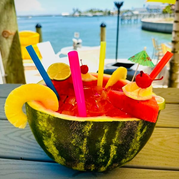 A watermelon cocktail with colorful straws and fruit garnishes sits on a table overlooking the water.