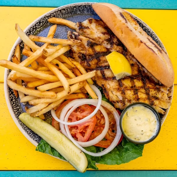 Grilled fish sandwich with fries, lettuce, tomato, onion, pickle, and tartar sauce on a colorful table.