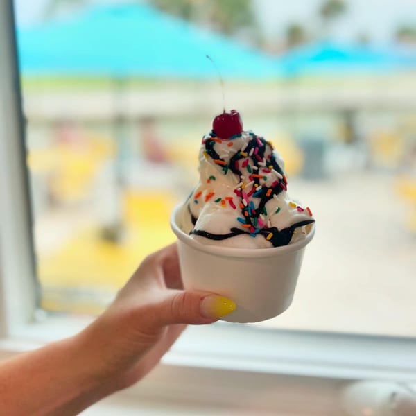 A hand holds a cup of vanilla ice cream with chocolate syrup, sprinkles, and a cherry on top.