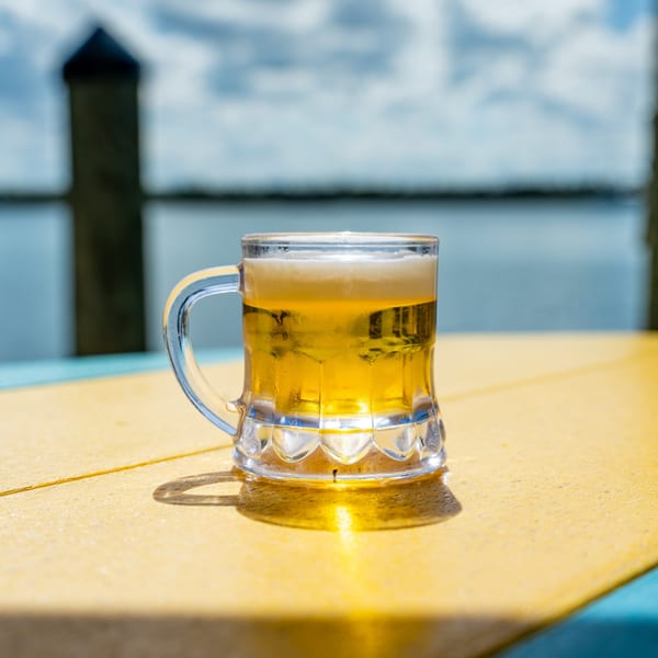 A frosty mug of beer sits on a yellow table with a body of water in the background.